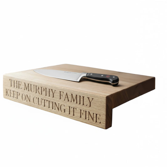 Personalised oak overhang chopping board engraved with ‘The Murphy Family Keep on Cutting It Fine,’ shown on a white background with chef’s knife resting flat on top.