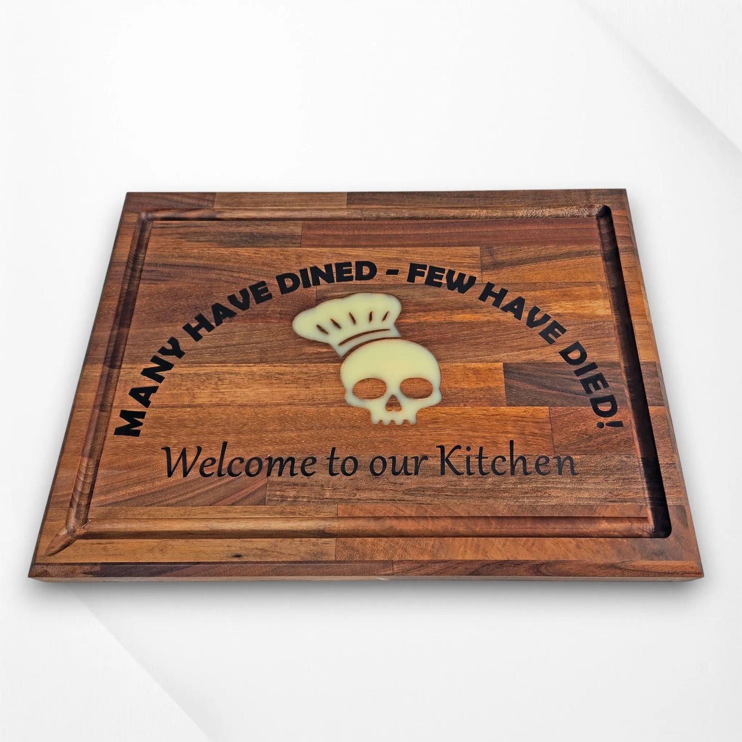 Funny engraved wooden chopping board with skull chef hat design and the phrase ‘Many Have Dined – Few Have Died! Welcome to our Kitchen,’ displayed on white background