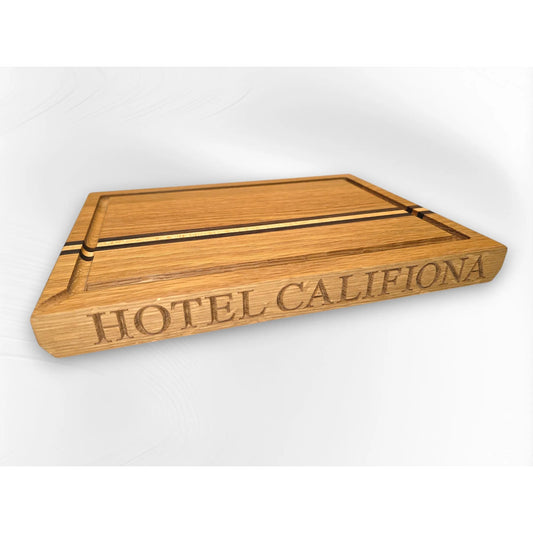 Personalised oak chopping board engraved with “HOTEL CALIFIONA” on the front edge, featuring inlaid stripes and a juice groove. Custom handmade wooden chopping board by The Crafty Oak with walnut and maple accent pieces.