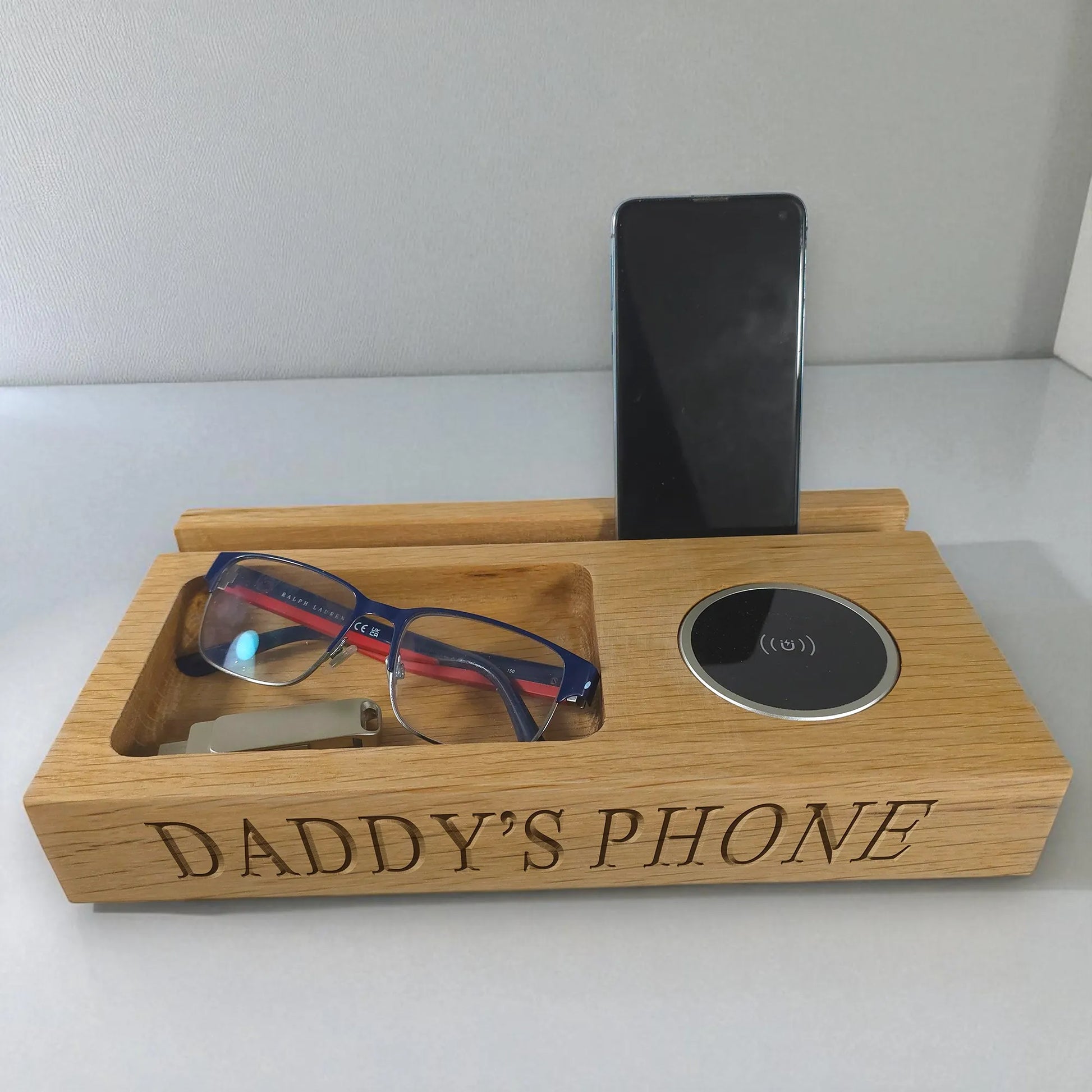 Wooden phone stand with engraved 'DADDY'S PHONE' holding a phone, glasses, and a USB drive.