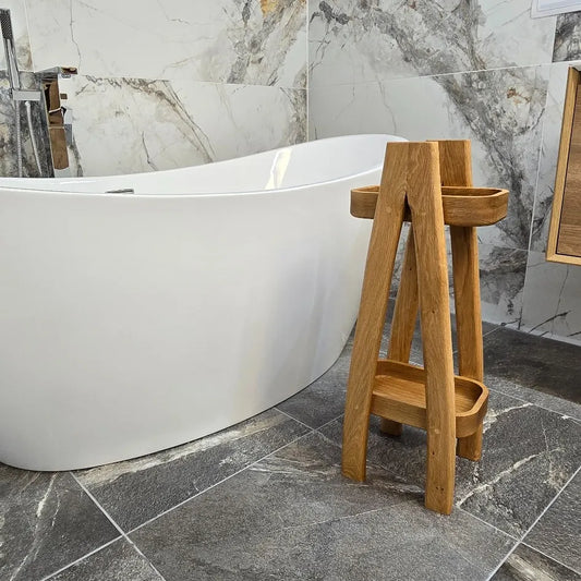 Modern freestanding oak bath caddy with tiered trays beside a luxury white bathtub in a marble-tiled bathroom. Handmade wooden bathroom organiser by The Crafty Oak.