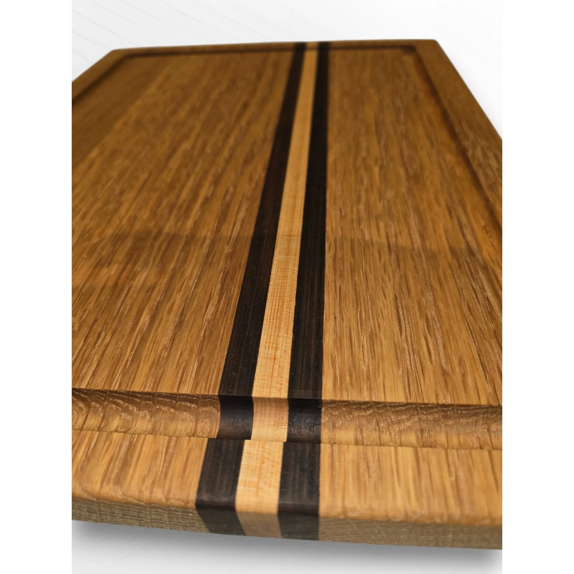 Close-up of handmade oak chopping board with contrasting walnut and maple inlay stripes, showing detailed wood grain and juice groove. The Crafty Oak premium wooden carving board.