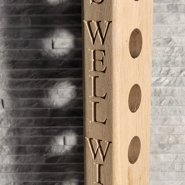 Close-up of vertical engraving on oak wine rack, showing carved ‘DRINKS WELL WITH OTHERS’ lettering and round bottle holes in solid wood against a stone tile background