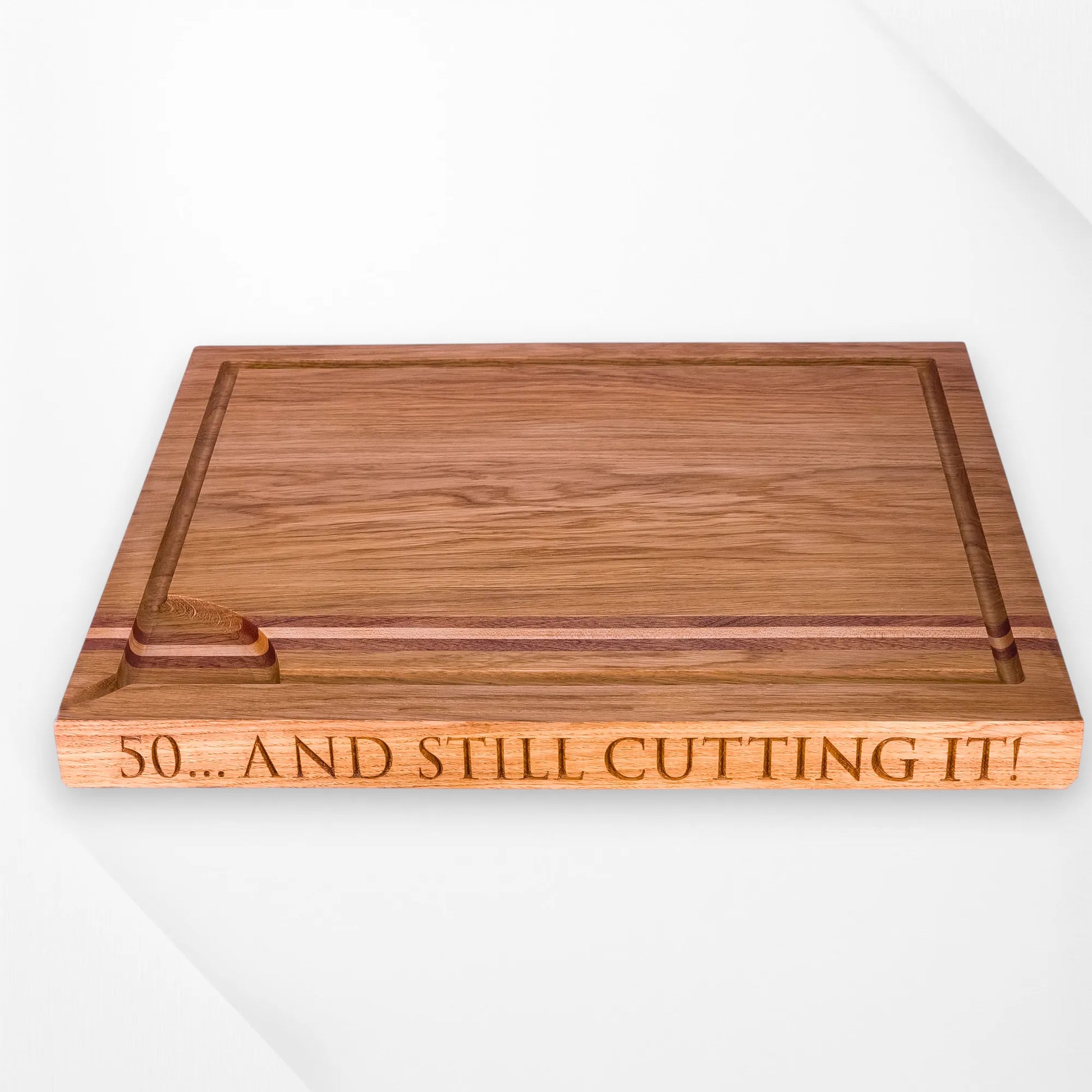 Personalised oak chopping board with engraved message "50... And Still Cutting It!" featuring juice groove and decorative strip inlay