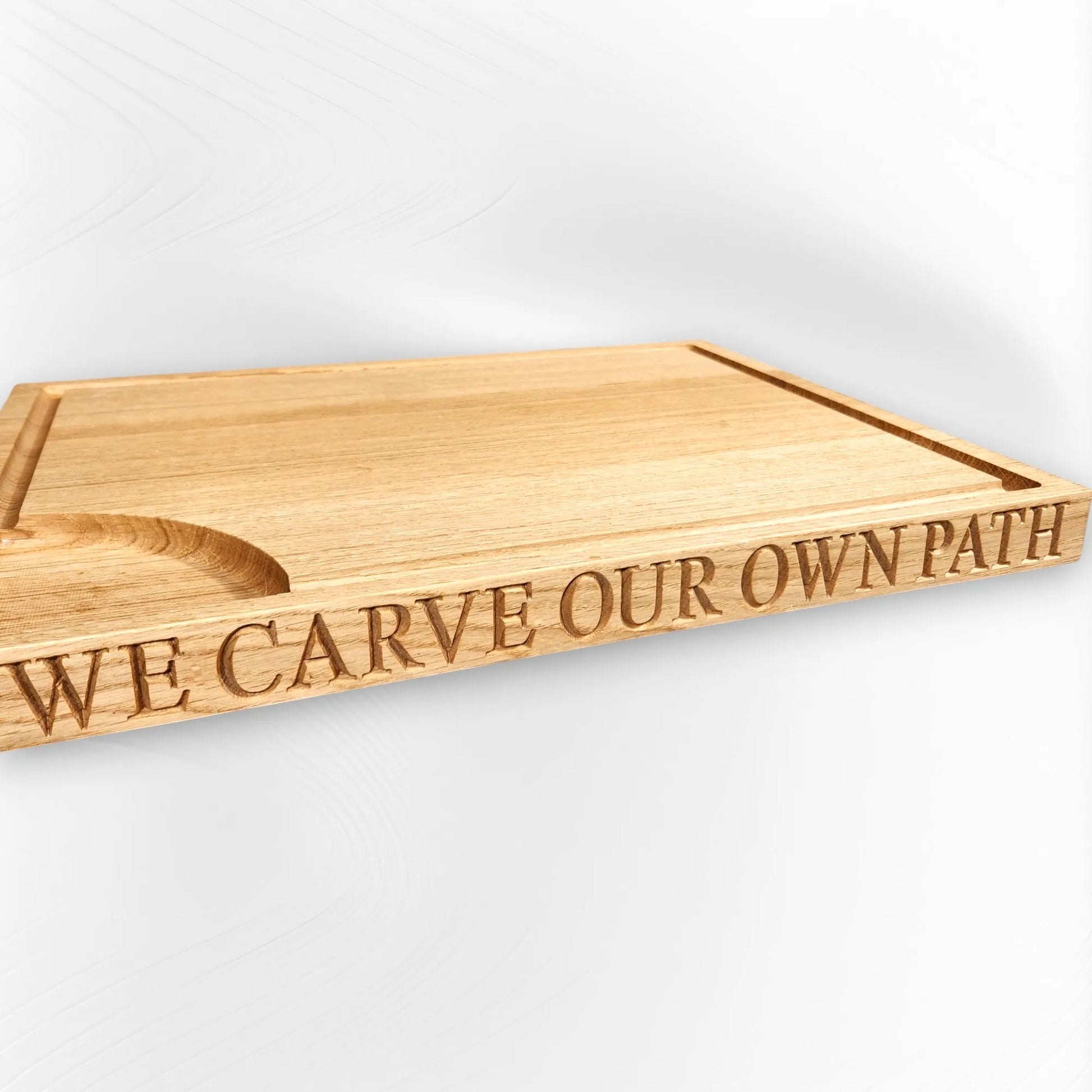 Angled view of personalised oak chopping board engraved with “WE CARVE OUR OWN PATH,” featuring a juice groove and utensil rest. Handmade wooden board by The Crafty Oak.