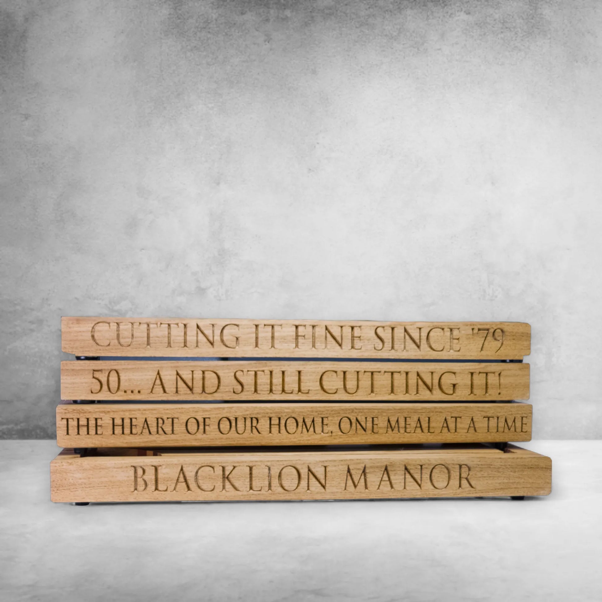 Stack of personalised oak chopping boards with engraved messages including "Cutting it fine since '79", "50... And Still Cutting It!", "The heart of our home, one meal at a time", and "Blacklion Manor"