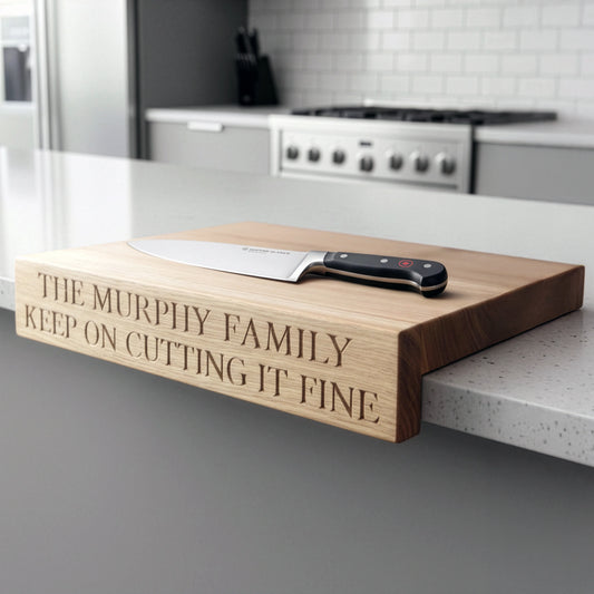 “Personalised oak overhang chopping board engraved with ‘The Murphy Family Keep on Cutting It Fine,’ shown on a modern kitchen worktop with chef’s knife resting flat on top.”