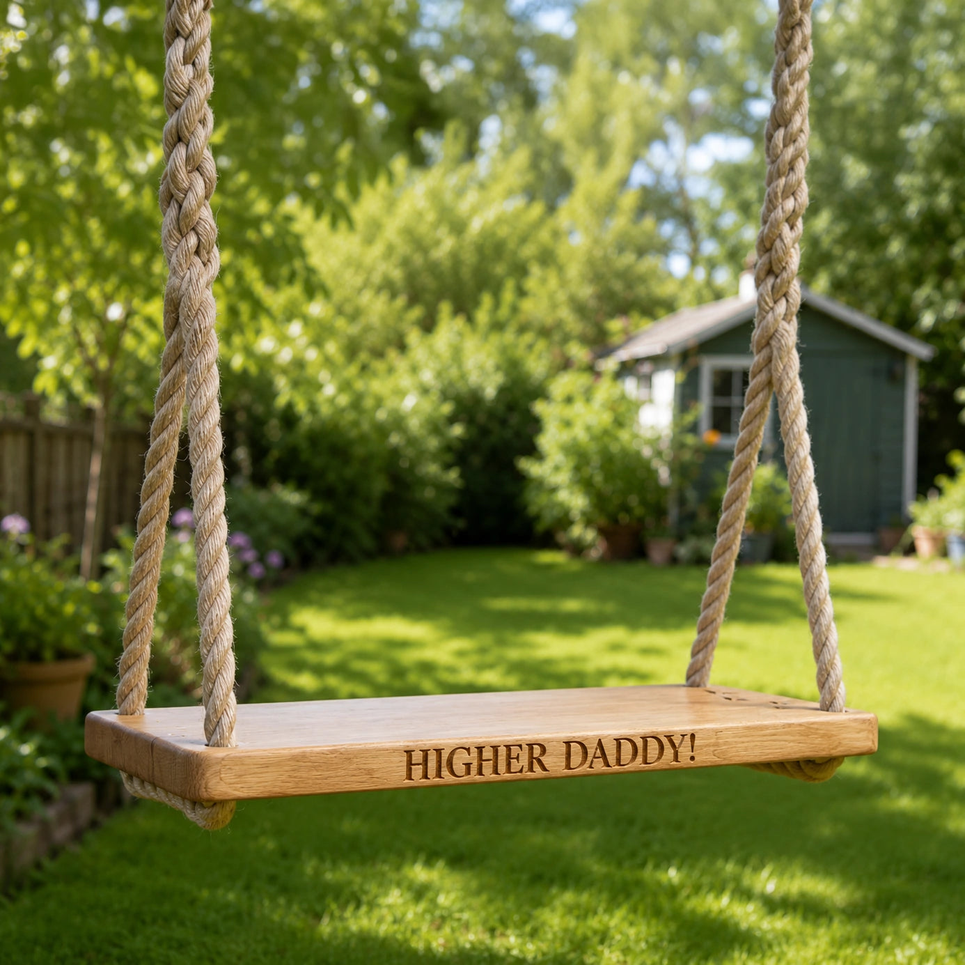 Personalised solid oak rope swing engraved with “Higher Daddy!” hanging in a sunny garden with a lawn and shed in the background.