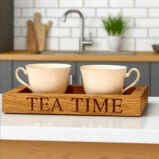 A pair of cream-coloured teacups sit inside a handcrafted oak tea tray engraved with the words "TEA TIME". The tray rests on a white countertop in a modern kitchen with a tiled backsplash and wooden worktop.