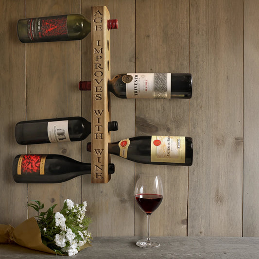 Wall-mounted personalised oak wine rack engraved with "Age Improves With Wine," holding six bottles of red wine horizontally. Set against rustic wood paneling with a glass of red wine and bouquet of white flowers on the counter below.