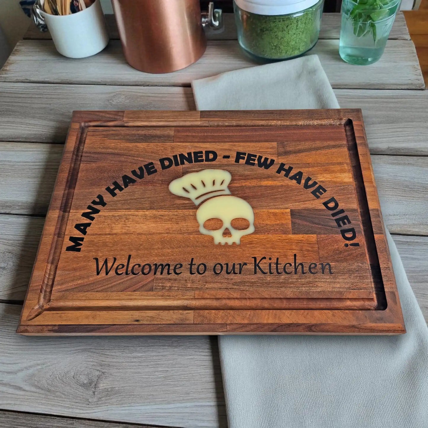 Funny engraved wooden chopping board with skull chef hat design and the phrase ‘Many Have Dined – Few Have Died! Welcome to our Kitchen,’ displayed on a rustic oak countertop with kitchen linens