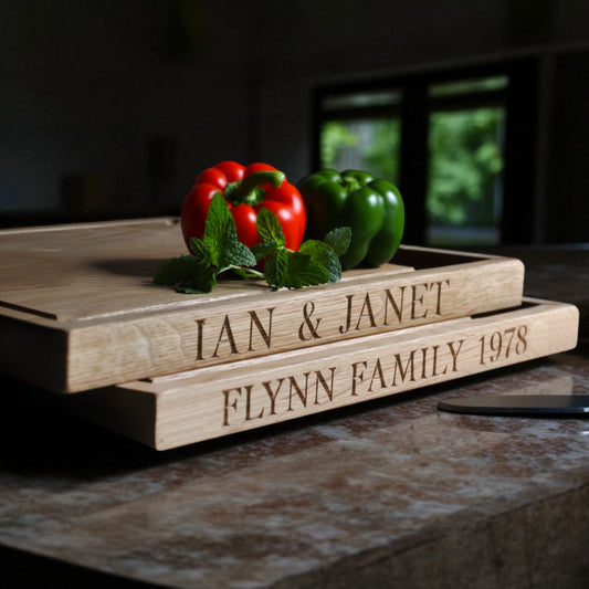 Two personalised oak chopping boards with engraved messages "Ian and Janet" and "Flynn Family" featuring juice groove and well. On a worktop with red green peppers.